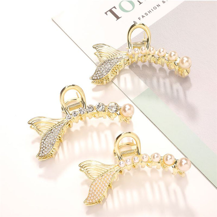 The SOHO Umi Hair Claw by SOHO features three gold mermaid tail-shaped clips with pearls and rhinestones, arranged on a white surface beside a magazine—perfect for creating magical, dreamy hairstyles.